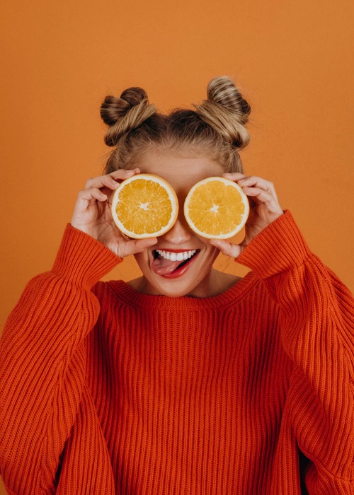 person in a bright orange sweater, holding two halves of an orange in front of their eyes, with a goofy smile