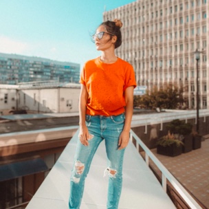 person wearing suglasses, orange t-shirt amd ripped jeans, looking to side off rooftop with buildings in the background, 